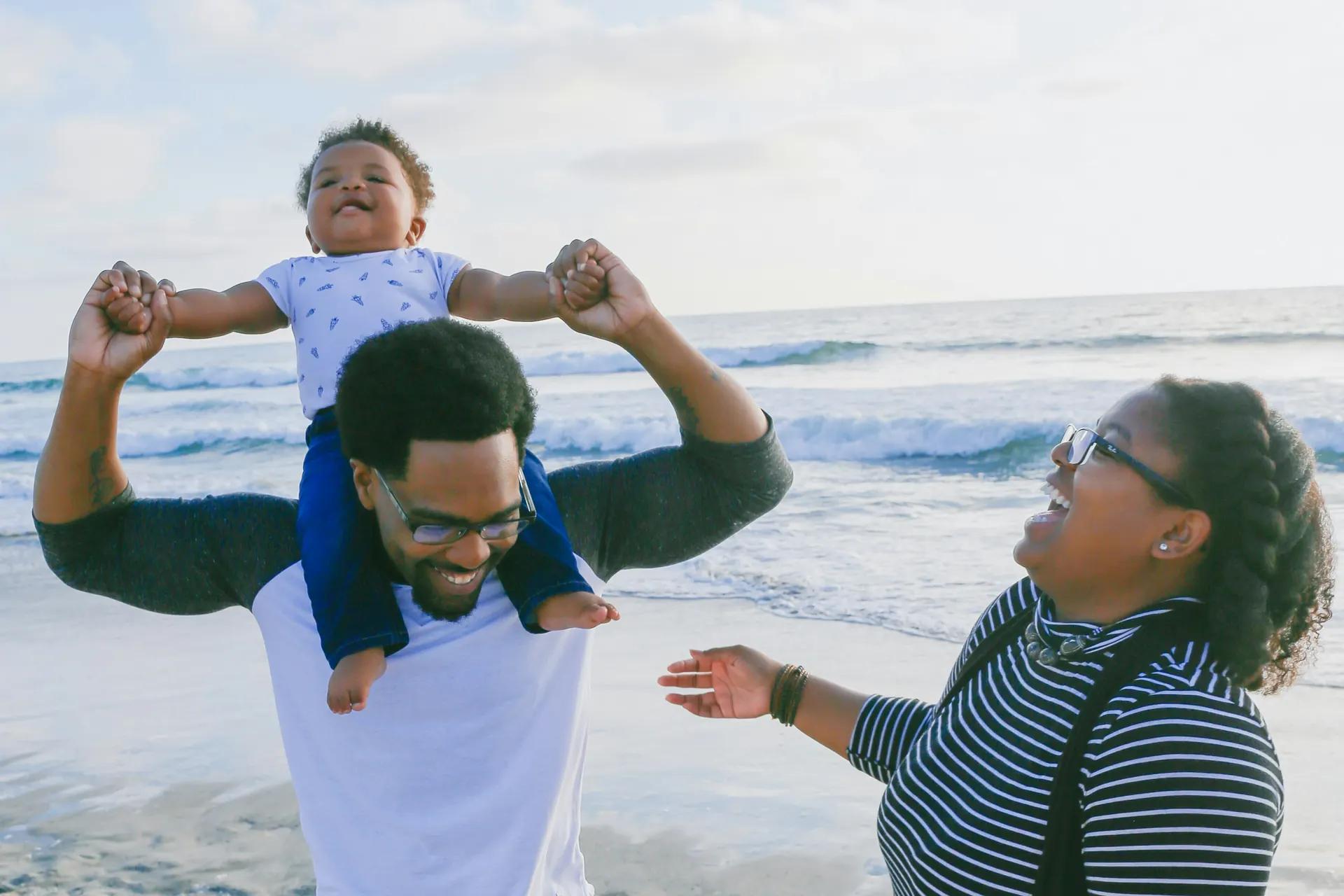 Un père porte son bébé sur les épaules, la mère rit à côté d’eux, au bord de la mer